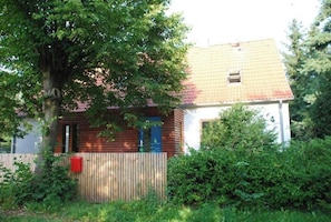 A wooden cabin with a red roof, surrounded by greenery and a wooden fence.