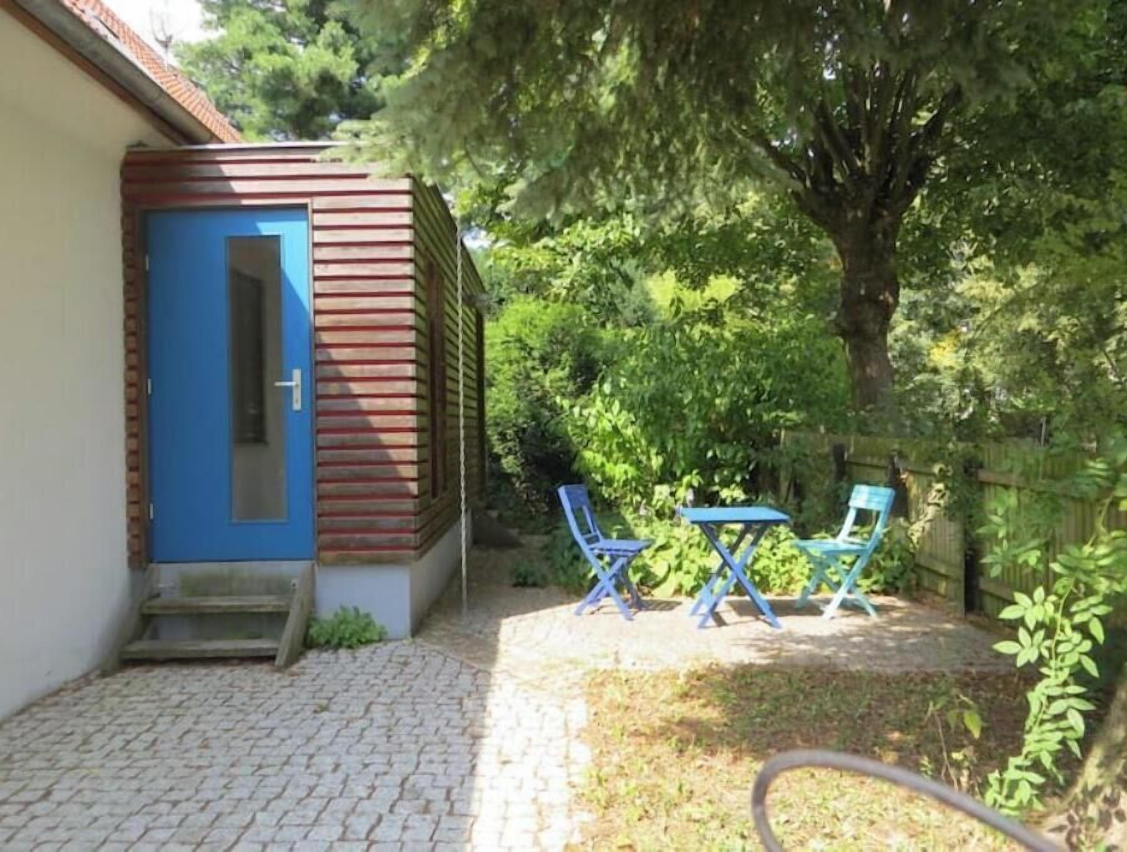 Ein kleines Gebäude mit einer blauen Tür, einer Ziegelaußenwand und einer gepflasterten Terrasse mit zwei blauen Stühlen.