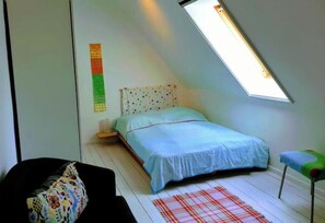 Une chambre avec un plafond en pente, un lit recouvert d’une courtepointe bleue, un coussin coloré, un tapis à carreaux et une petite chaise verte.