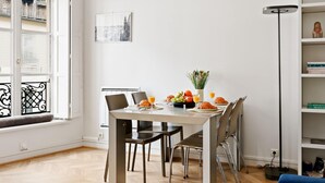 A dining area with a table set for two, a window with blinds, a bookshelf, and a floor lamp. - Stylish 1BR in the Heart of Paris by Louvre (Paris)