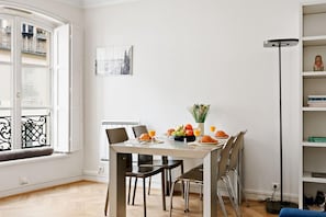A dining area with a table set for two, a window with blinds, a bookshelf, and a floor lamp. - Stylish 1BR in the Heart of Paris by Louvre (Paris)