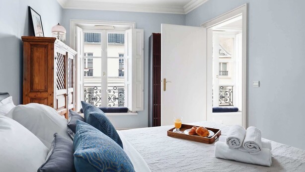 A bedroom with a bed, pillows, a tray with croissants and juice, and a view of balconies through the window. - Stylish 1BR in the Heart of Paris by Louvre (Paris)