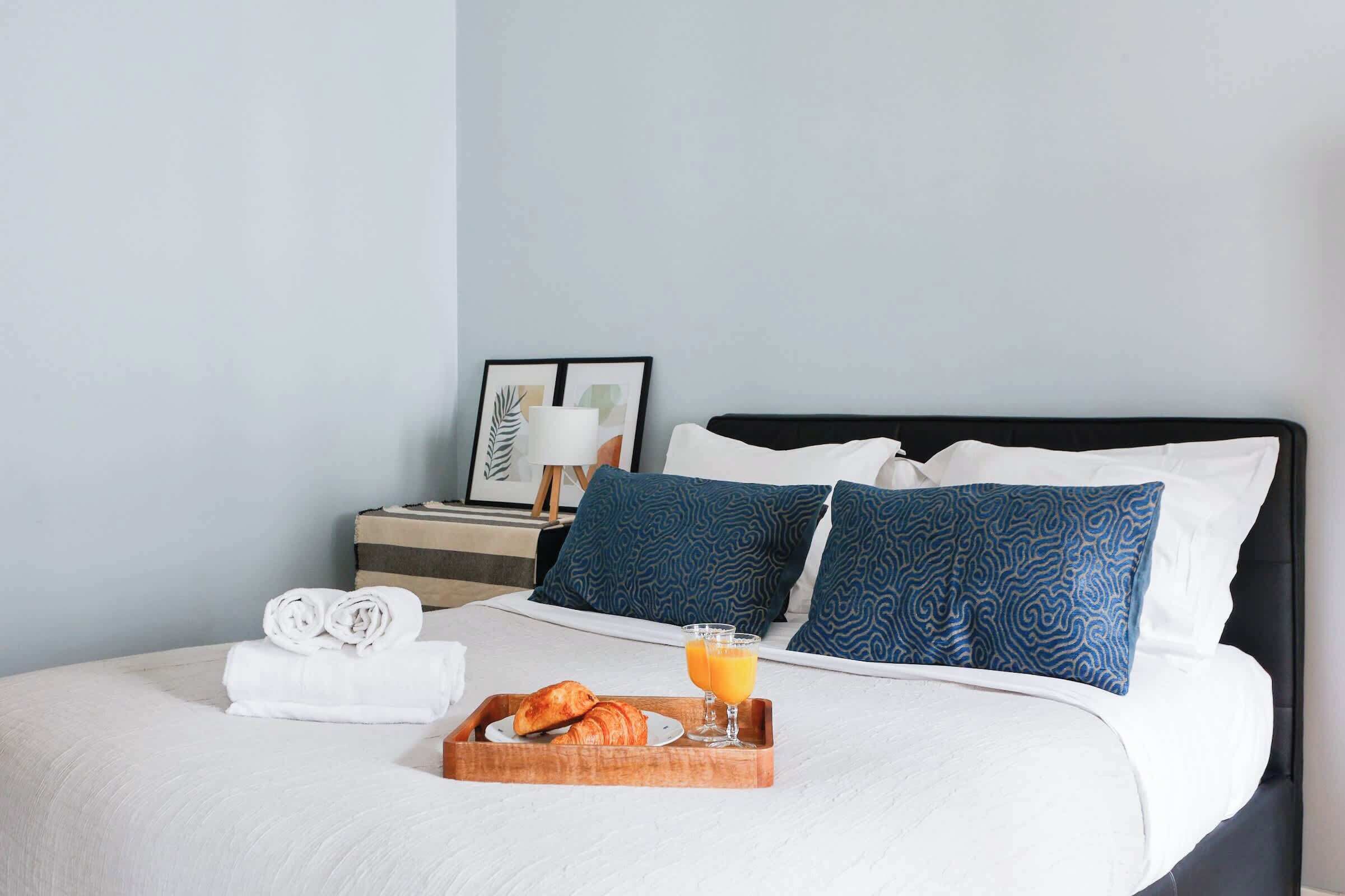Een keurig opgemaakt bed met blauw gebloemde kussens, een dienblad met croissants en sinaasappelsap, en een nachtkastje met een lamp en een lijst met kunstwerk.