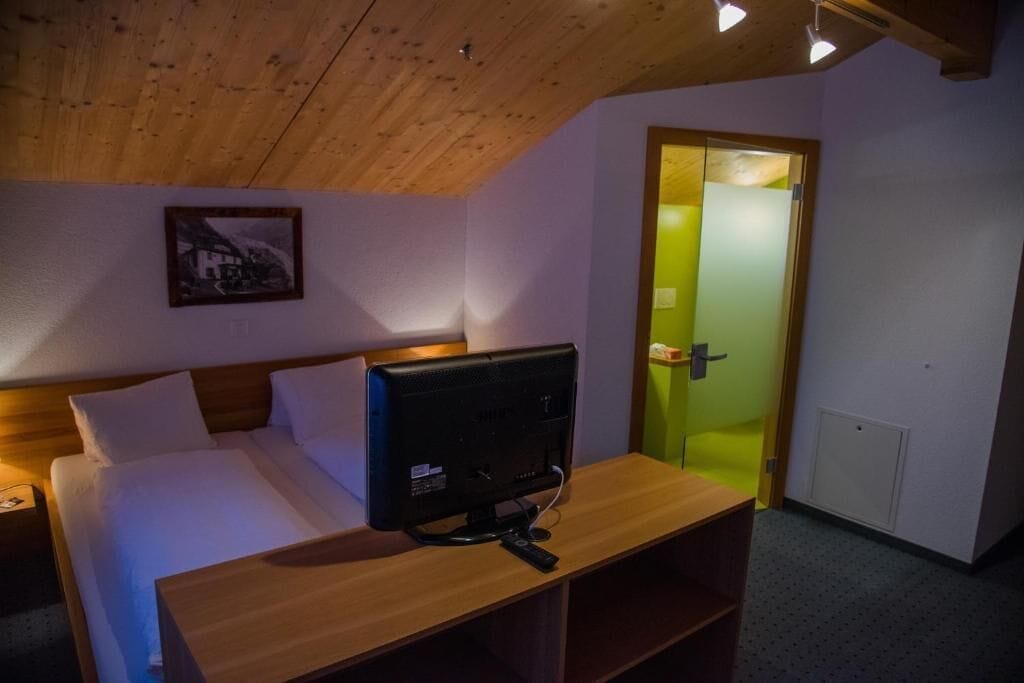 Un dormitorio con cama, un televisor sobre una mesa de madera, un cuadro en la pared y una puerta iluminada de verde.