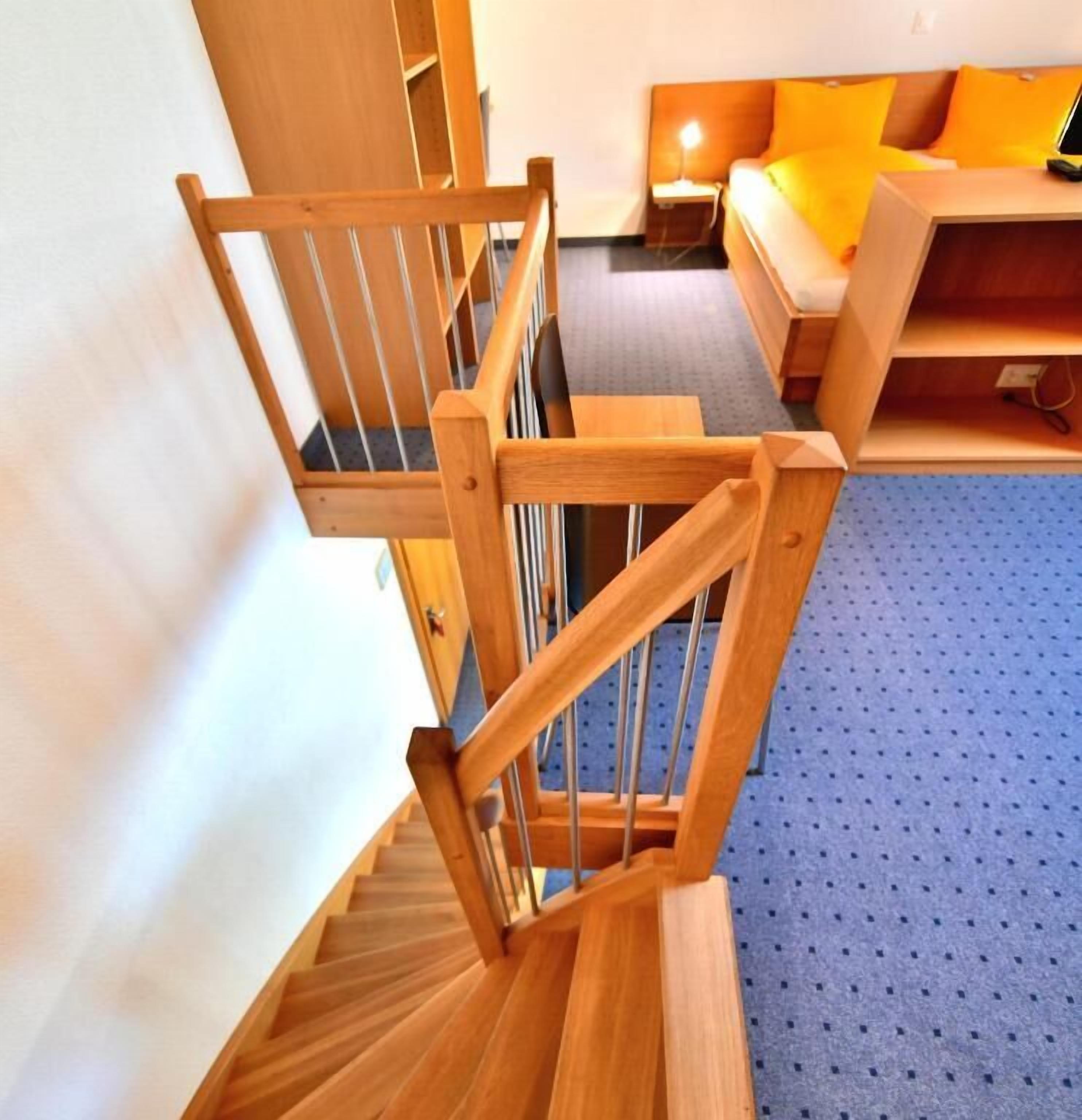 Un escalier en bois, dont les marches sont recouvertes d’une moquette bleue, mène à une pièce où se trouvent un lit et une table de chevet.