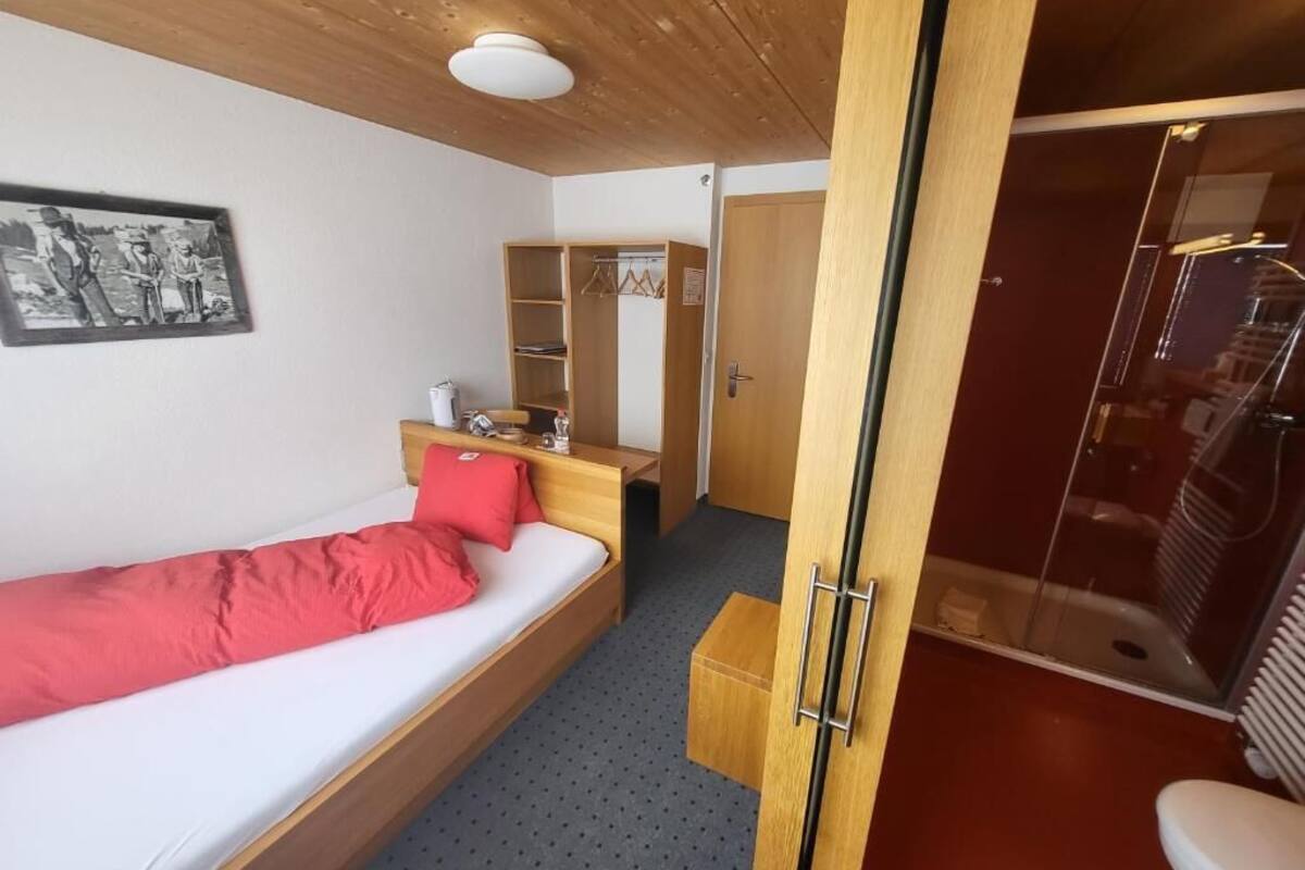 A single bed with a red blanket, a wooden bedside table, a mirror, a wardrobe, and a shower area.