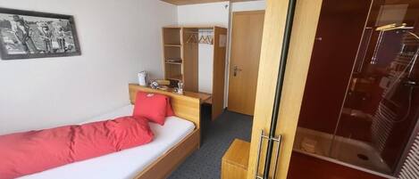 A single bed with a red blanket, a wooden bedside table, a mirror, a wardrobe, and a shower area.