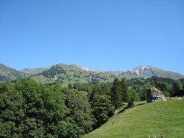 Paisagem montanhosa sob um céu limpo, com um primeiro plano gramado e um pequeno edifício.
