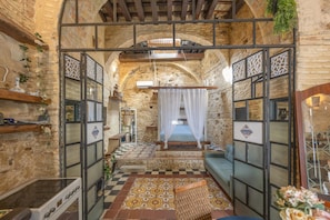 A stone-walled room with a checkered floor, a bed with white curtains, and a small table with a vase.