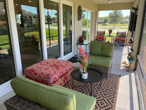 A sunlit patio with a green sofa, a patterned ottoman, and a small table with a vase of flowers.