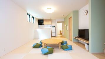 A modern living room with a round table, green and blue cushions, a flat-screen TV, and a kitchen in the background.