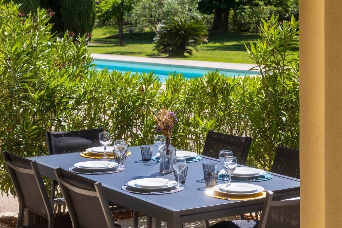 An outdoor dining area with a table set for six, surrounded by lush greenery and a view of a swimming pool.