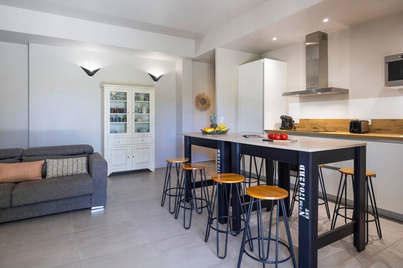 Cocina moderna con una isla central, taburetes de bar, horno empotrado y un armario blanco con puertas de cristal.