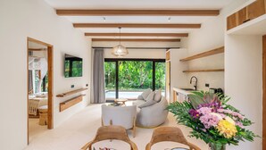 A modern living room with a sofa, chairs, a dining table, a TV, and a view of a garden. - Hutan Villa - Open Bath and Private Pool (Ubud)