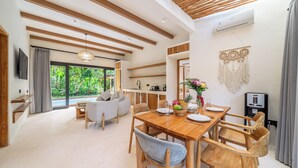 A modern interior with a wooden dining table, chairs, a sofa, and a kitchen area. - Hutan Villa - Open Bath and Private Pool (Ubud)