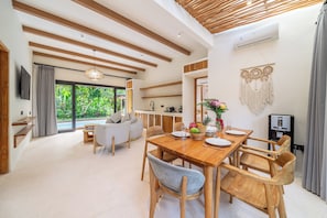 A modern interior with a wooden dining table, chairs, a sofa, and a kitchen area. - Hutan Villa - Open Bath and Private Pool (Ubud)