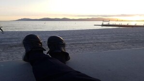 Beine einer Person, die sich an einer Kante über Wasser ausruhen, mit Blick auf einen Pier und Berge in der Ferne.