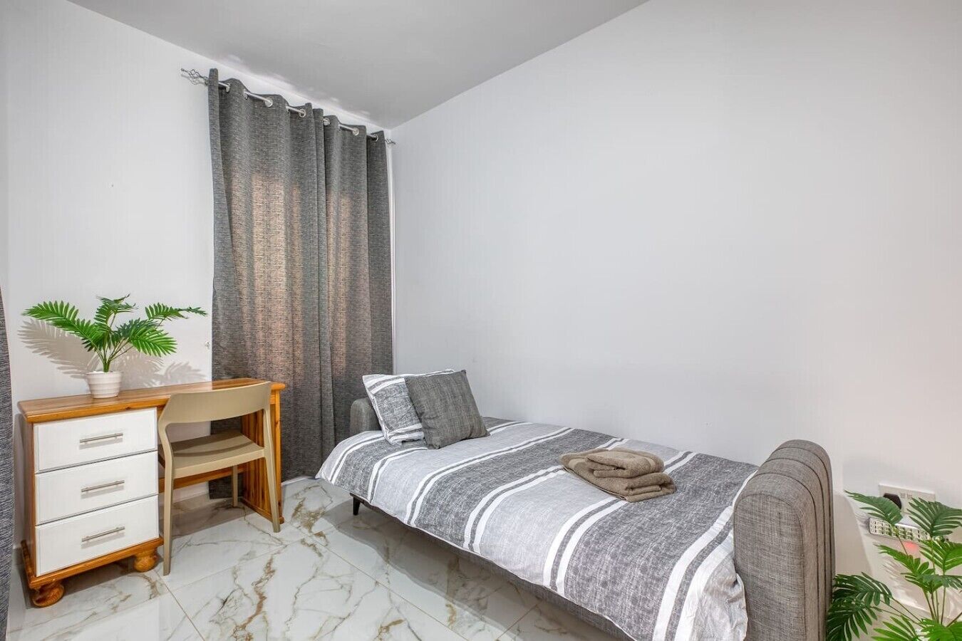 Un dormitorio con cama, escritorio, silla, planta en maceta y cortina.