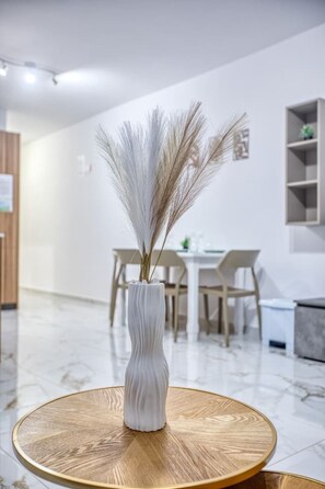 Ein modernes Interieur mit einer weißen Vase auf einem runden Holztisch, einem Essbereich mit Stühlen und einer weißen Wand mit einem Bild.