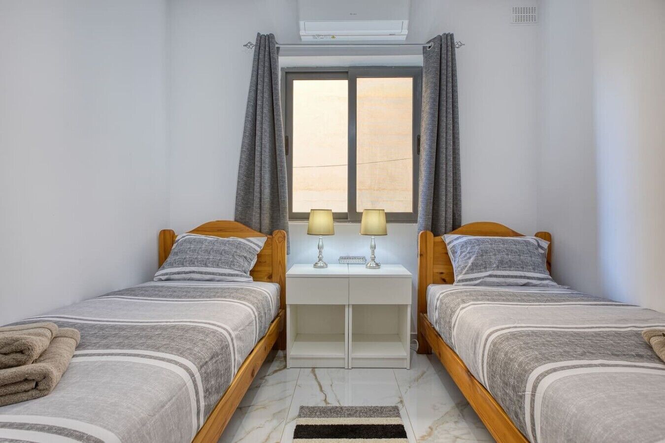 A twin bedroom with two beds, a small desk with lamps, a window with curtains, and an air conditioning unit.