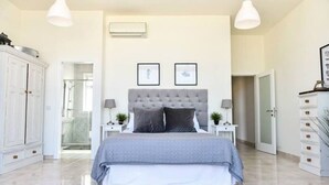 Un dormitorio con una cama grande, dos mesitas de noche, un armario y una puerta que da a otra habitación.