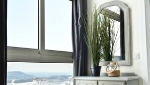 Una habitación con vista a edificios, una ventana con cortina, un espejo, un aparador y una planta en maceta.