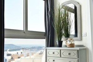 A room with a view of buildings, a window with a curtain, a mirror, a dresser, and a potted plant. - Luxury Maltese Penthouse with Breathtaking views (Il-Mellieħa)