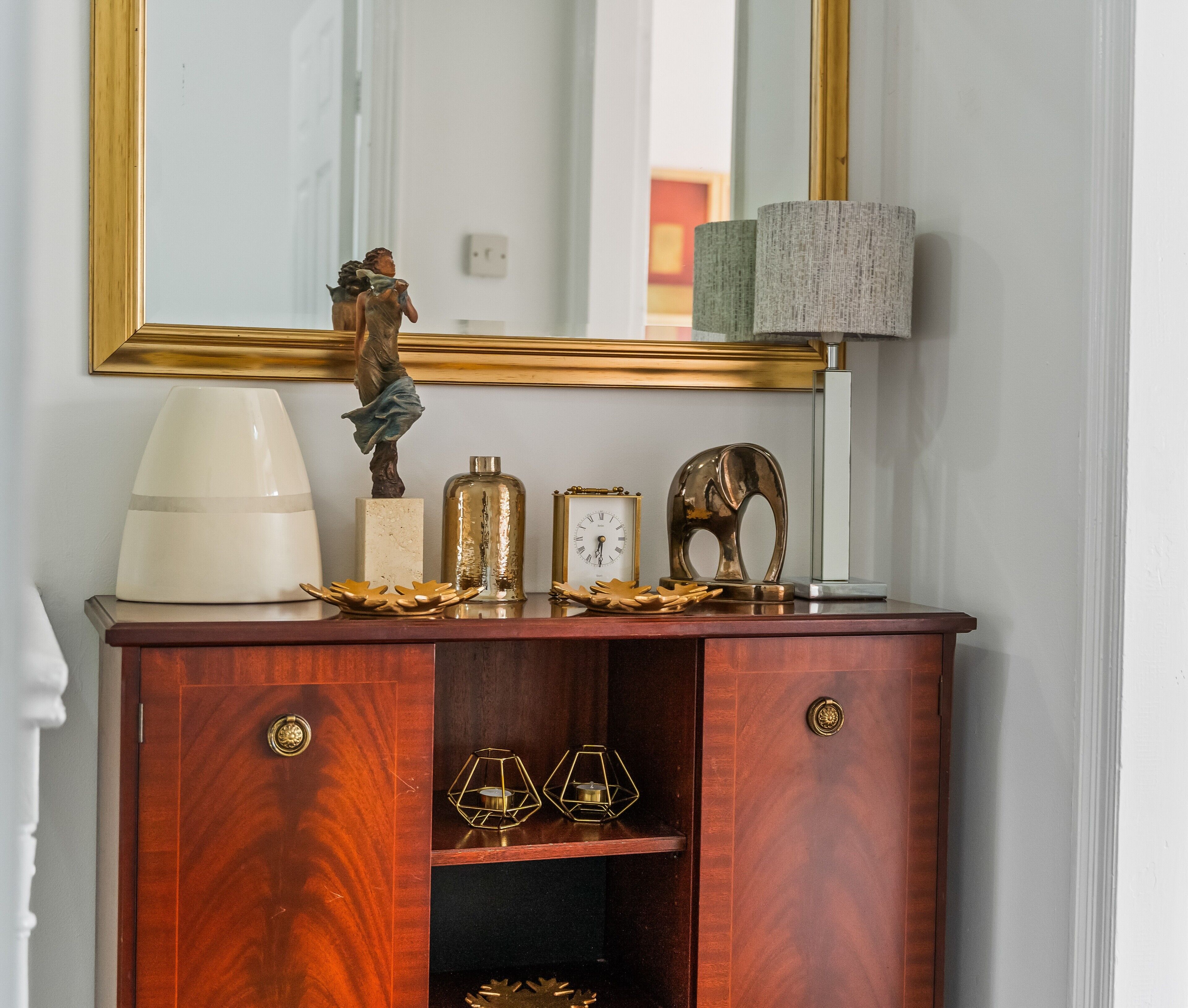 Une commode en bois, agrémentée d’un miroir, d’une lampe et d’objets décoratifs, notamment une horloge et une sculpture.
