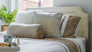 Un lit bien fait, avec une tête de lit, une plante en pot et un plateau sur lequel se trouvent un croissant et une tasse.