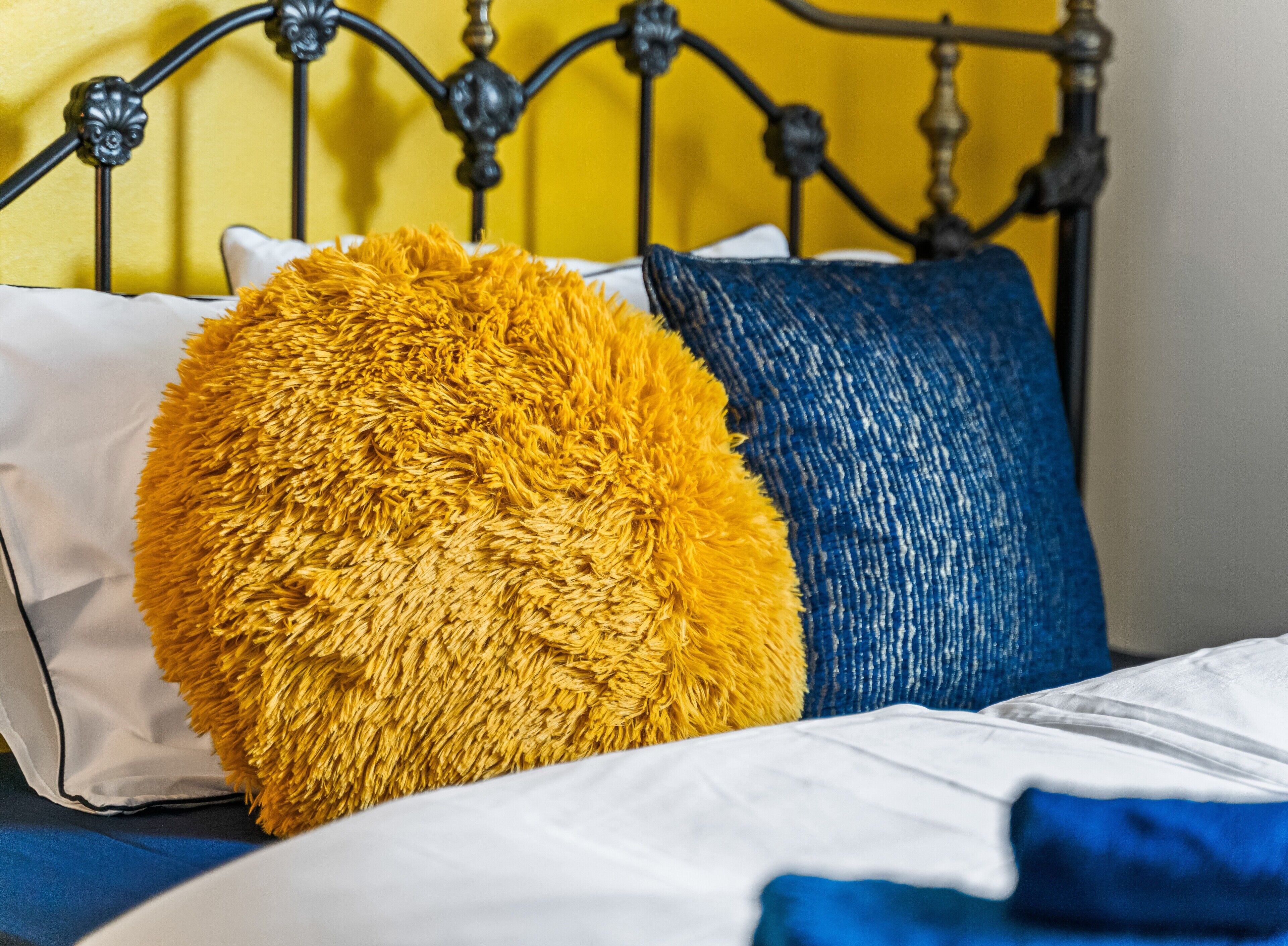 A bed with a metal headboard, a white pillow, a large fluffy yellow pillow, and a blue pillow.