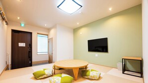 A room with tatami flooring, a round wooden table, green cushions, a television on the wall, and a door with a handle. - Irodori Hotel KAZE (fukuokashi,hakataku,toukou)