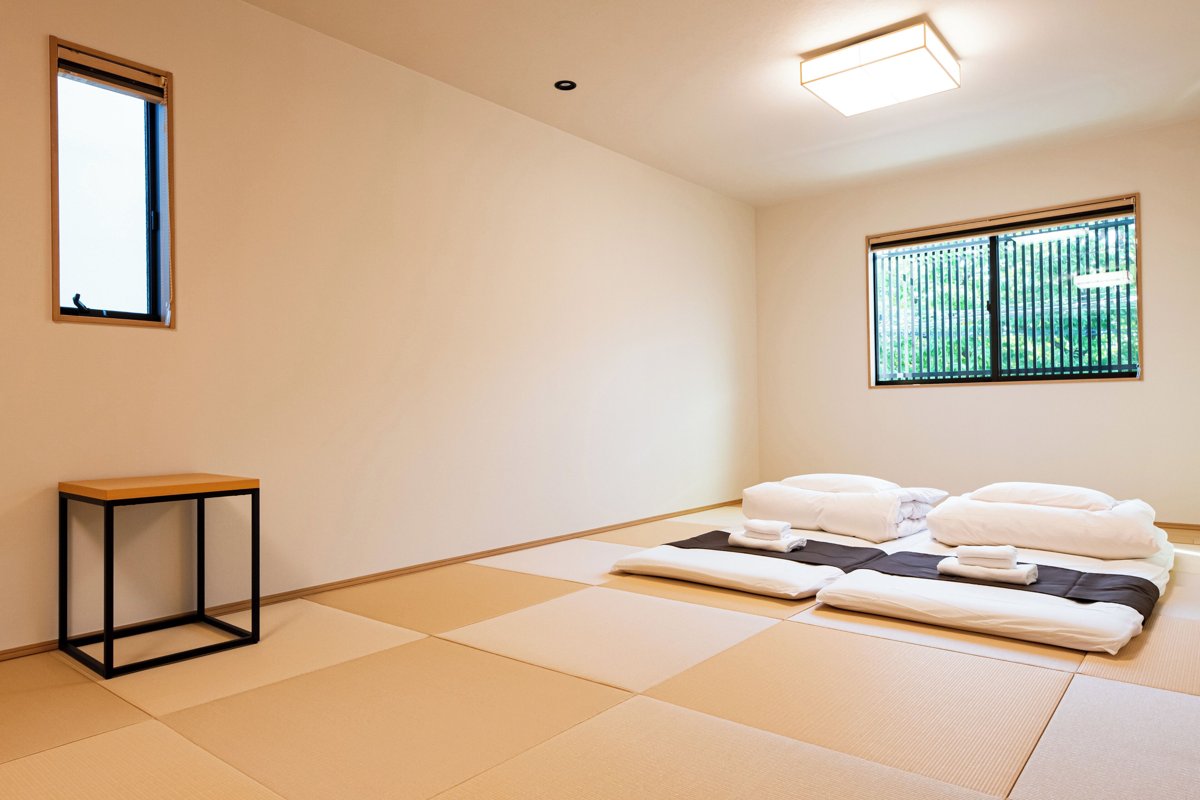 A room with tatami flooring, a bed with white bedding, a small table, and two windows.