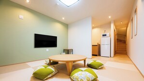 A modern living room with a round wooden table, green and patterned cushions, a flat-screen TV, and a kitchen area with a refrigerator and microwave. - Irodori Hotel KAZE (fukuokashi,hakataku,toukou)
