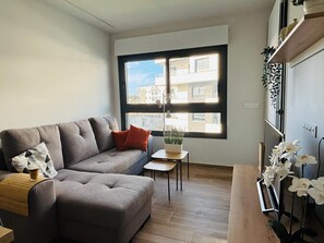 Ein modernes Wohnzimmer mit einem grauen Sofa, einem kleinen Tisch mit Pflanze und einem Fenster mit Blick auf Gebäude.