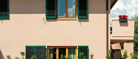 A building with a flat roof, two windows with green shutters, and a small balcony with red flowers.