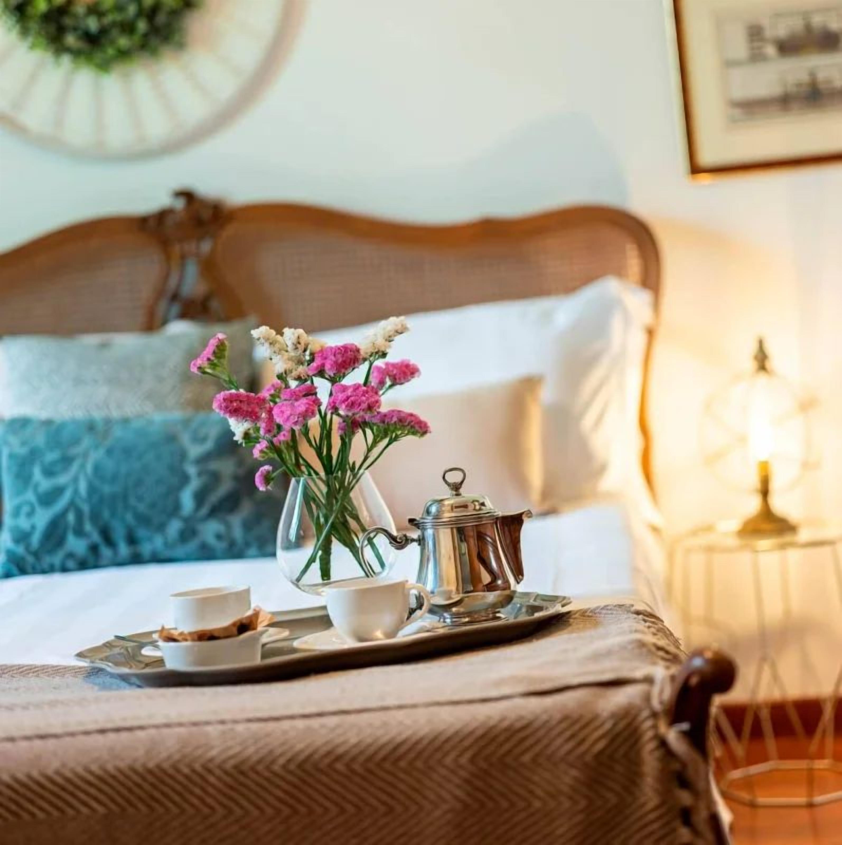 A neatly made bed with a headboard, a tray with a teapot, cups, and a small bowl of food, and a vase with flowers on a table.