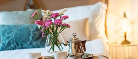 A neatly made bed with a headboard, a tray with a teapot, cups, and a small bowl of food, and a vase with flowers on a table.