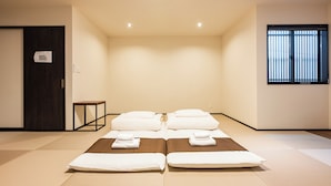 Una habitación con esteras de tatami, dos camas con sábanas blancas, una mesita, una puerta de madera oscura y una ventana con persianas.