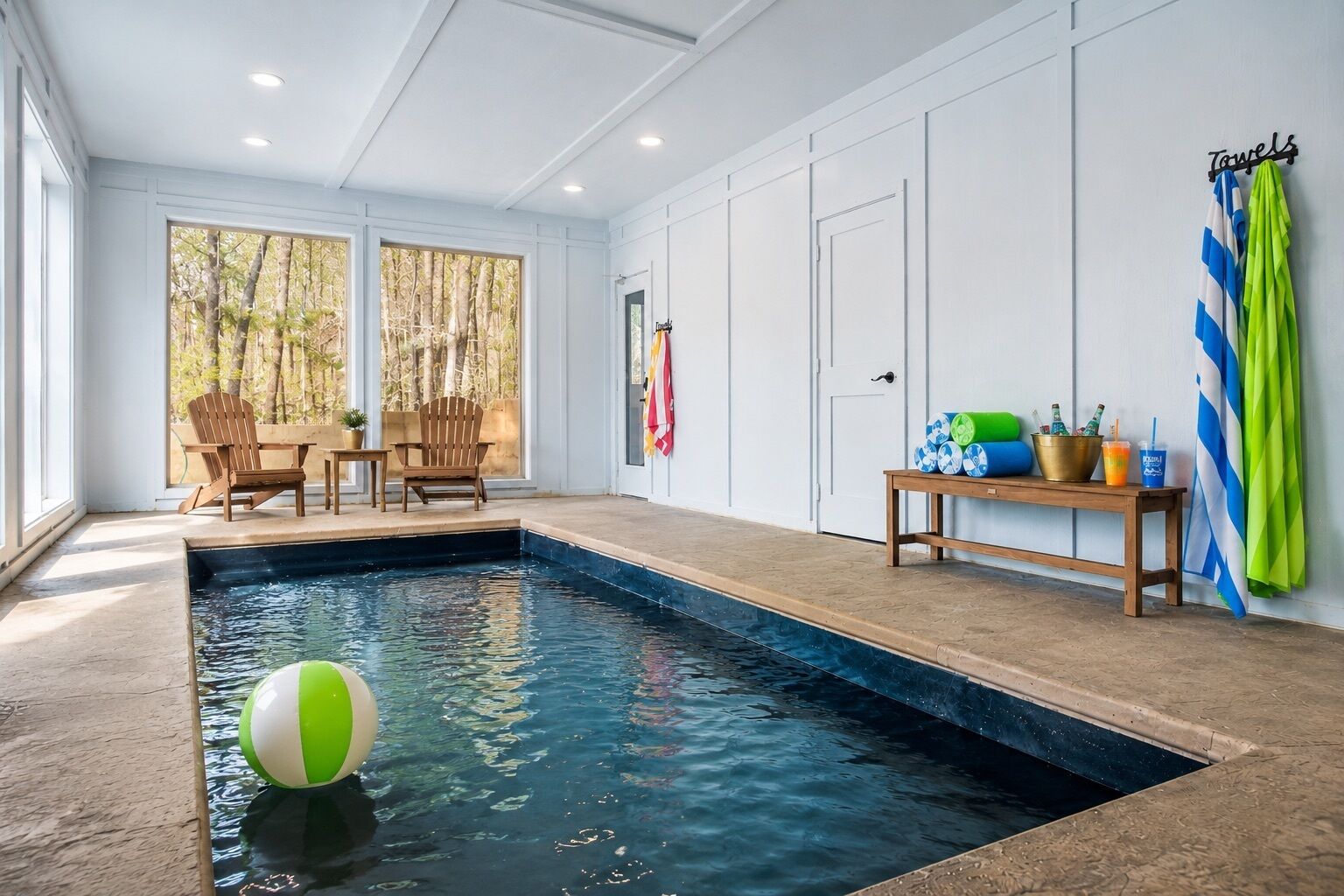 A swimming pool with a wooden deck, two chairs, a table with drinks, and a flag on the wall.