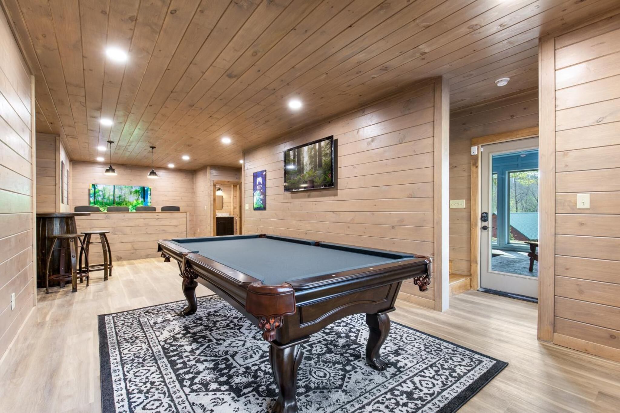 A pool table in a room with wooden walls and ceiling, a patterned rug, and a door leading outside.