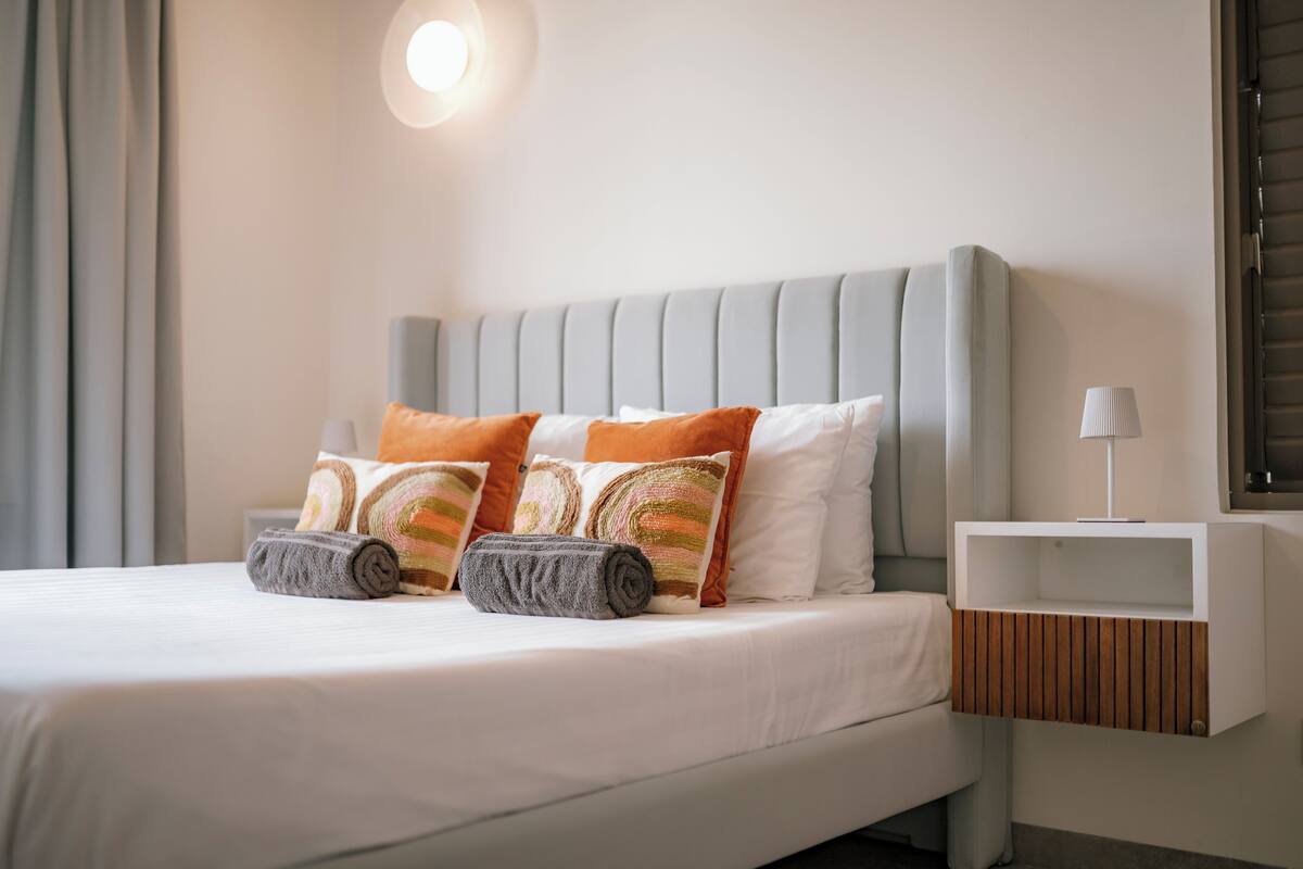 A neatly made bed with pillows and a rolled towel, a bedside table with a lamp, and a wall-mounted light fixture.