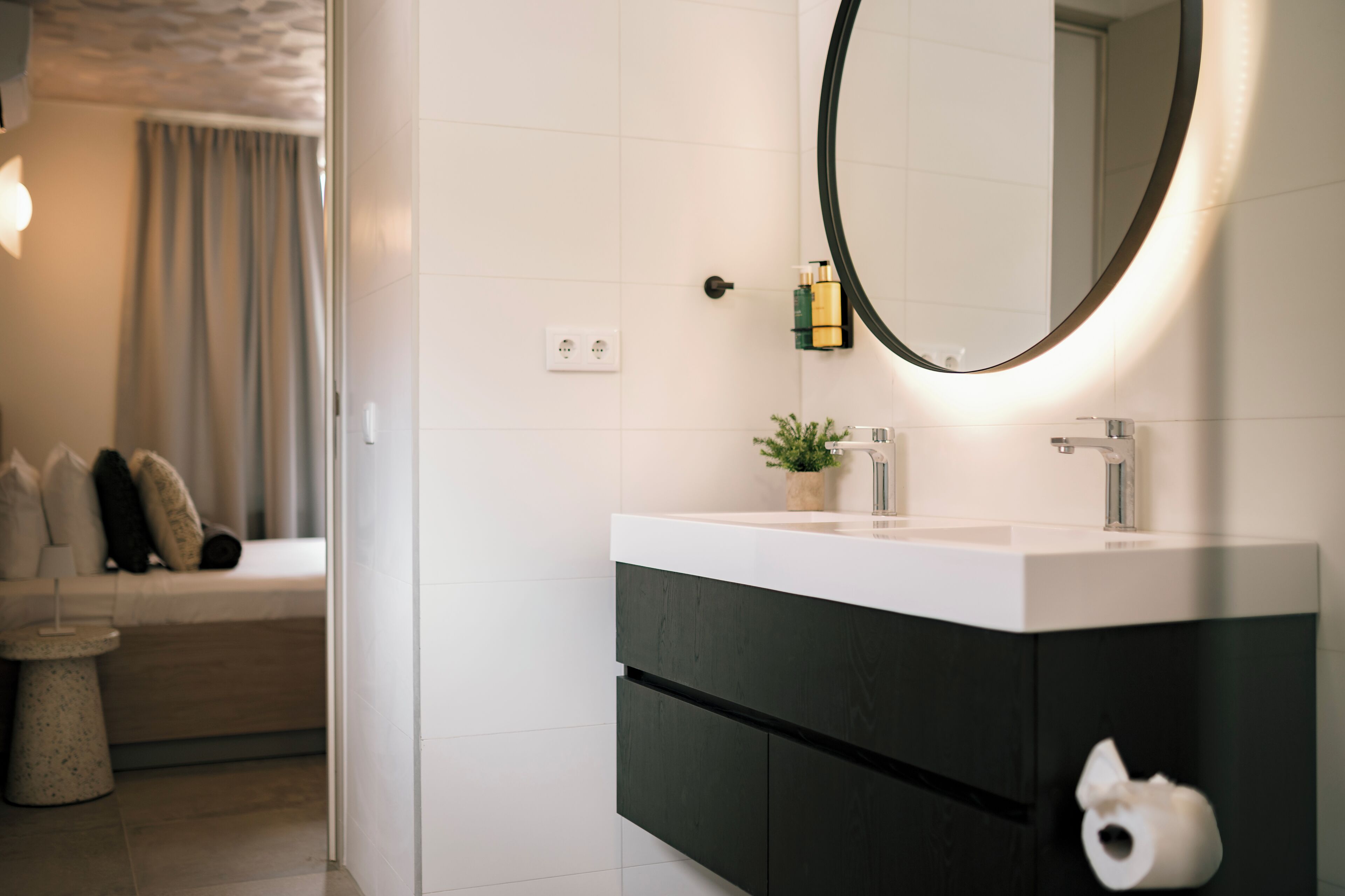 Un baño moderno con un lavamanos blanco, mueble oscuro y un espejo redondo con luz.