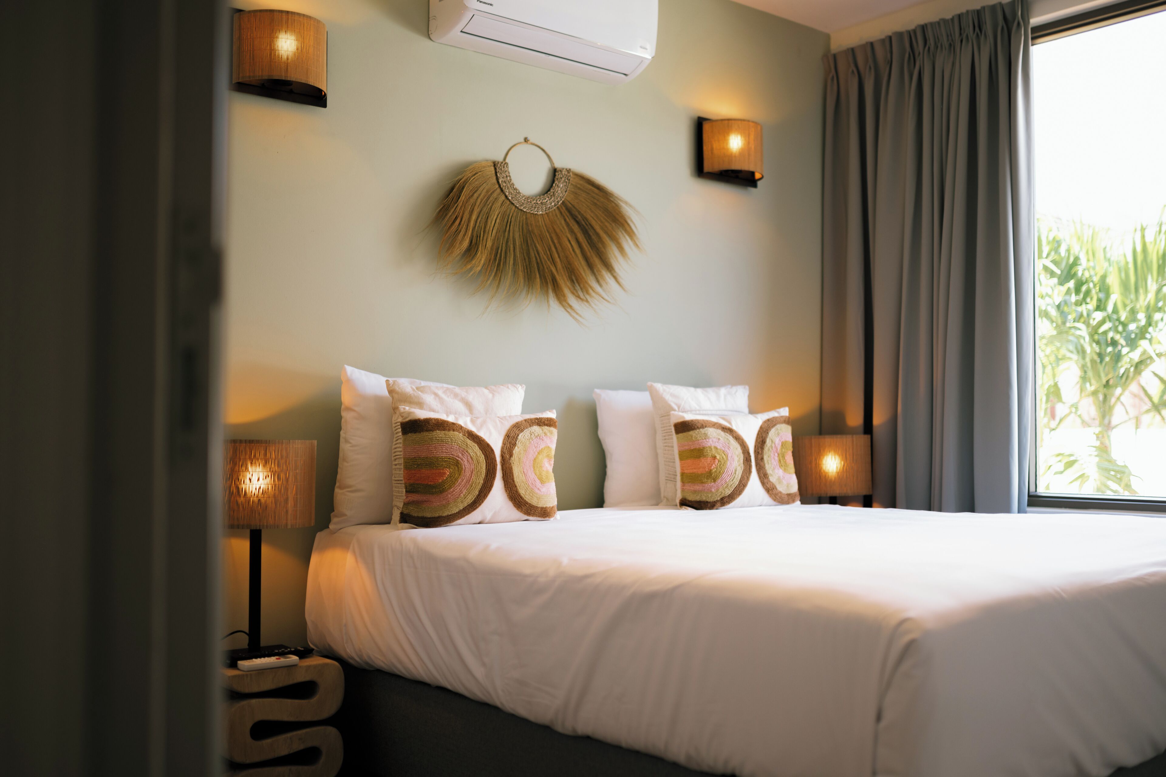 A hotel room with a bed, bedside lamps, a wall-mounted light fixture, and a window with curtains.