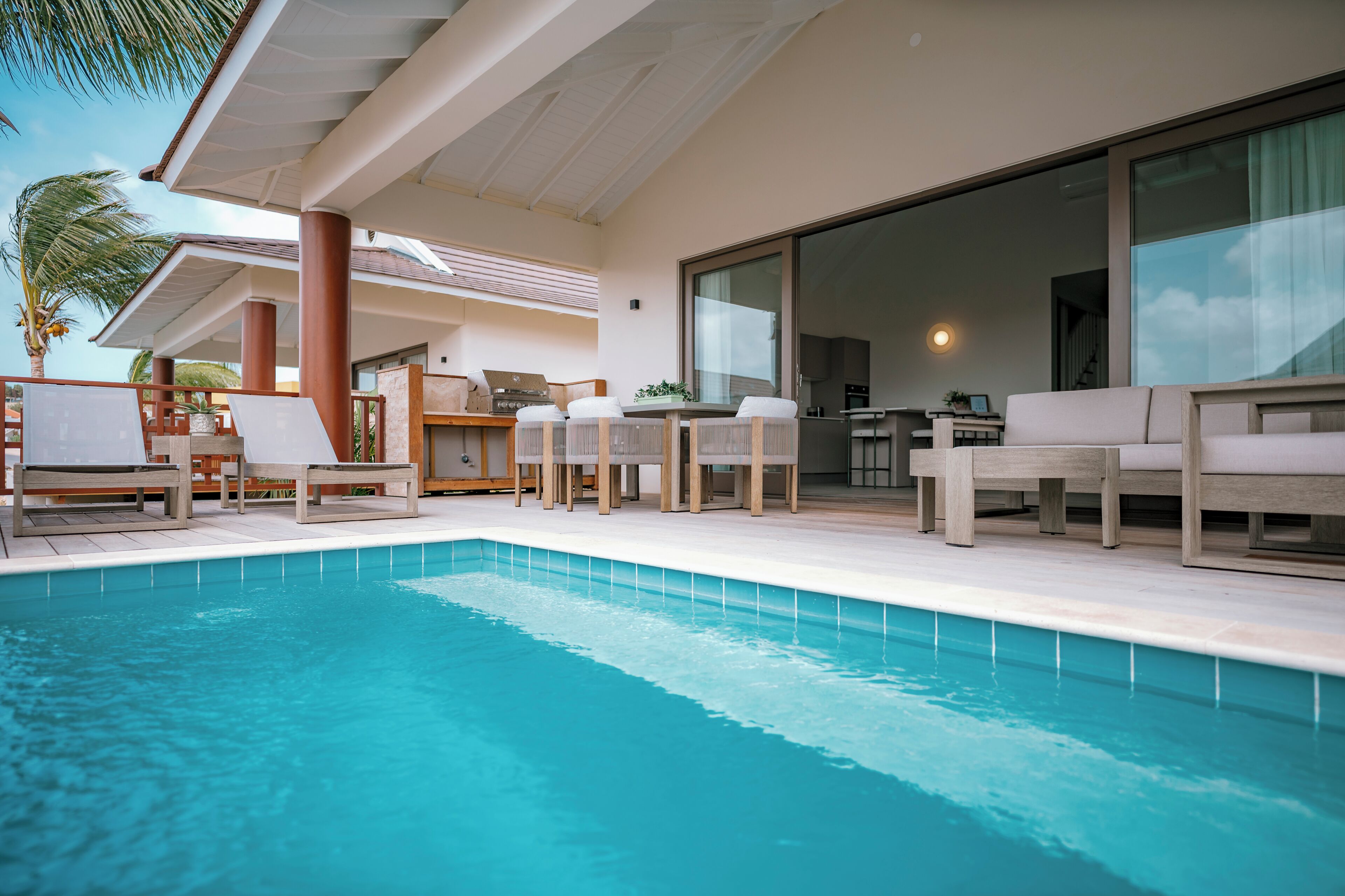 Zona junto a la piscina con sillones de descanso, una mesa de comedor y una cocina visible a través de un ventanal.