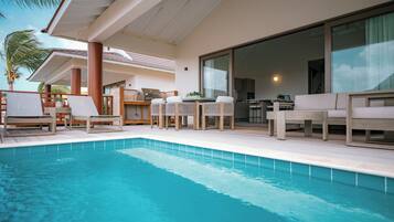 Zona junto a la piscina con sillones de descanso, una mesa de comedor y una cocina visible a través de un ventanal.