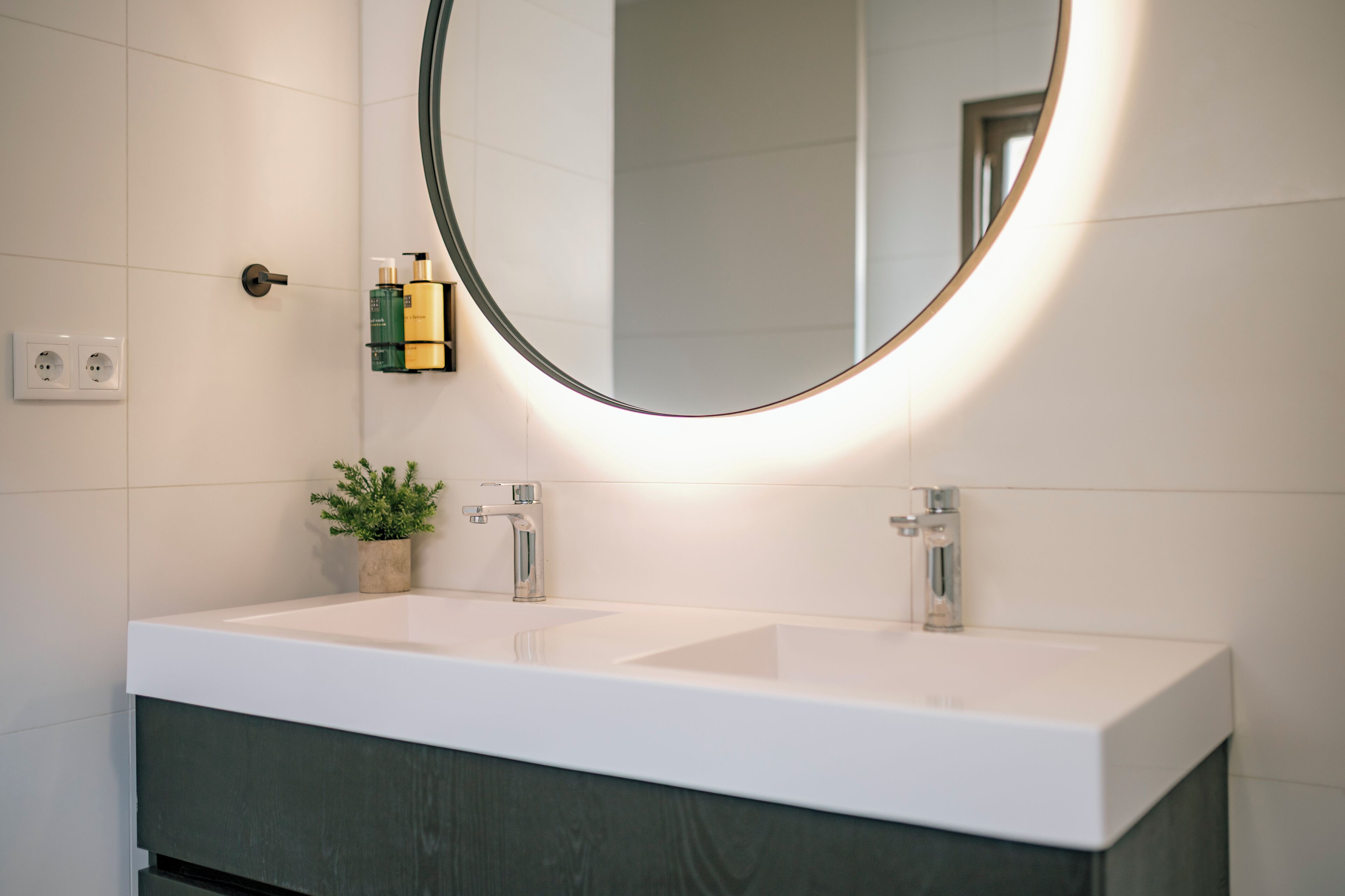 Un baño moderno con un lavamanos blanco, un espejo redondo y una planta en maceta.