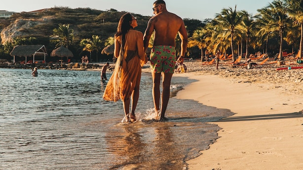 A beach scene with a couple walking along the shore, palm trees, and beach umbrellas. - The Jazmyn - Blue Bay Golf & Beach Resort (Sint Michiel)
