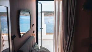A bedroom with a bed, a bedside table, a lamp, a mirror, and a view of the sea through a large window. - Experience a brand-new seafront duplex penthouse in Ghadira Bay, featuring 2 bedrooms, 2 bathrooms, a fully equipped kitchen and two spacious terraces, both offering beautiful sea views just steps from the beach. (Il-Mellieħa)