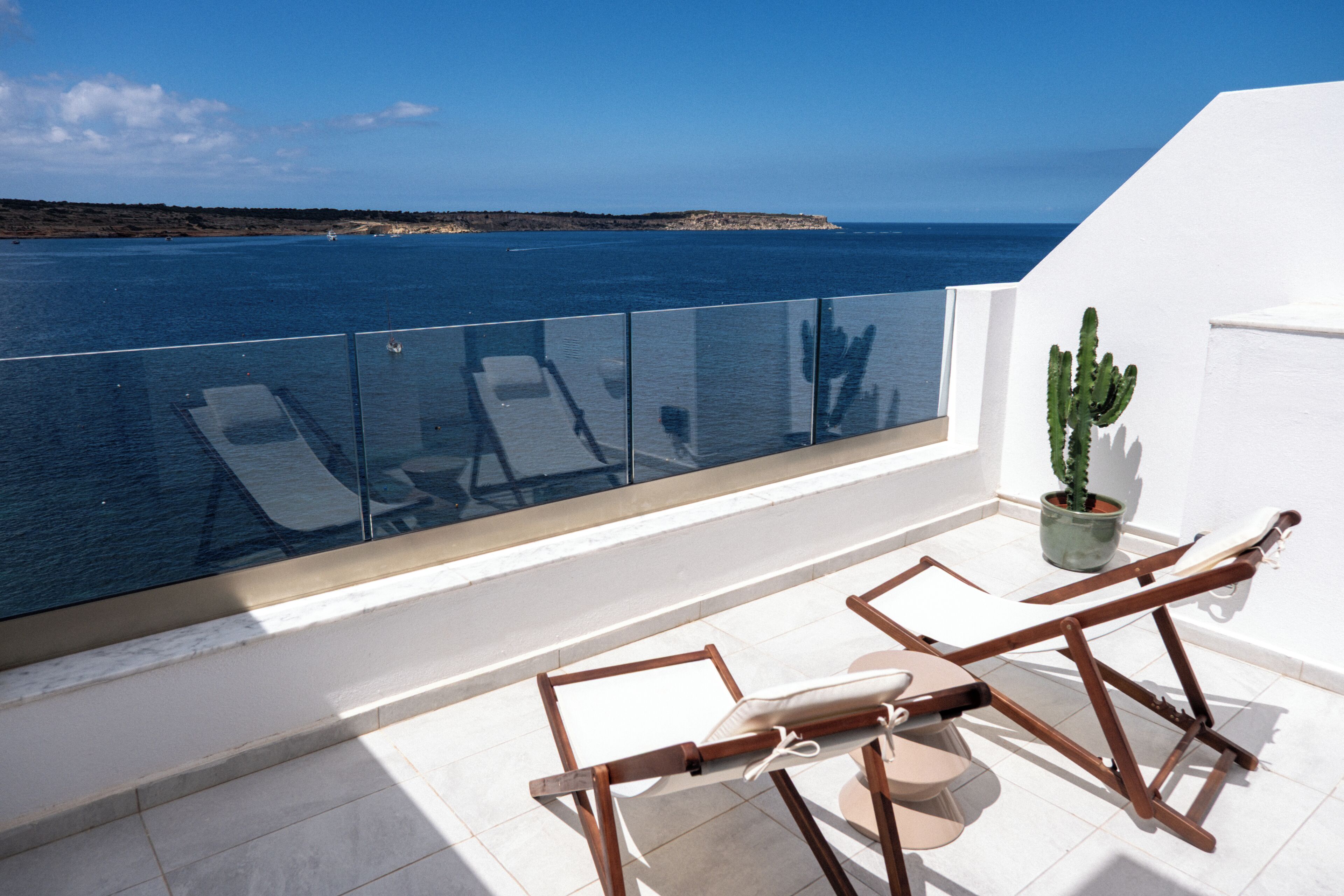 Ein Balkon mit Glasgeländer, Blick auf das Meer, eine Kakteepflanze und ein Holzstuhl.