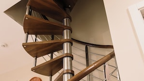 A spiral staircase with metal railings and wooden steps, a clock on the wall, and a window with blinds. - Experience a brand-new seafront duplex penthouse in Ghadira Bay, featuring 2 bedrooms, 2 bathrooms, a fully equipped kitchen and two spacious terraces, both offering beautiful sea views just steps from the beach. (Il-Mellieħa)